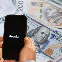 Dłoń trzyma smartfon z logo Revolut na tle różnych walut. To banki, które nie współpracują z komornikiem, oferując alternatywę dla tradycyjnych instytucji.