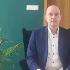 Mężczyzna w garniturze siedzi w fotelu obok rośliny, na tle zielonej ściany i drewnianych drzwi.