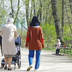 Kobieta z wózkiem spaceruje po parku, w tle inne osoby i dziecko na ławce.