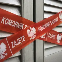Czerwona taśma z orłem i napisem "KOMORNIK SĄDOWY ZAJĘTE" krzyżuje się na tle szarej, żaluzjowej powierzchni. To znak, że nastąpiło zajęcie komornicze bez wezwania do zapłaty.