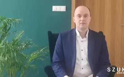 Mężczyzna w garniturze siedzi w fotelu obok rośliny, na tle zielonej ściany i drewnianych drzwi.