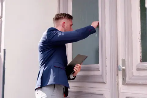Mężczyzna w garniturze puka do drzwi, trzymając teczkę. Czy zastanawiasz się, ile komornik może zabrać z L4?