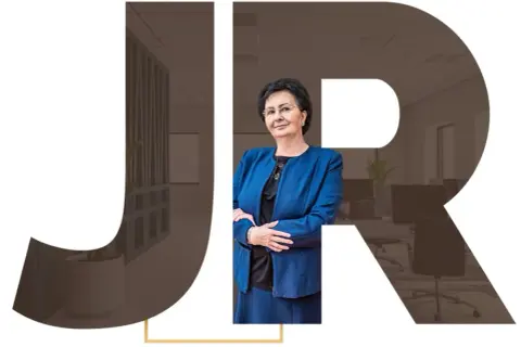 Kobieta w niebieskiej marynarce na tle liter JR i wnętrza biura.