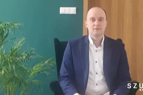 Mężczyzna w garniturze siedzi w fotelu obok rośliny, na tle zielonej ściany i drewnianych drzwi.
