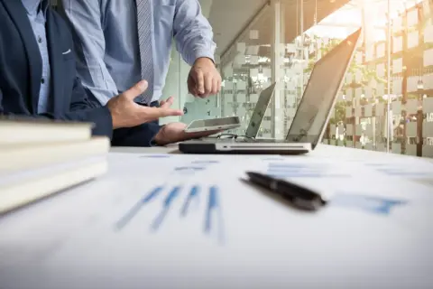 Dwóch mężczyzn w garniturach analizuje dokumenty i tablet w nowoczesnym biurze.