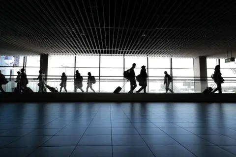 Sylwetki ludzi z bagażami na ruchomym chodniku lotniska, w tle okna z jasnym niebem. Długie kolejki i zatrzymania na lotnisku za długi to codzienność podróżnych.