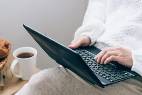Osoba w białym swetrze uczy się, jak czytać raport BIK, pracując na laptopie przy kubku kawy i koszyku z włóczką.