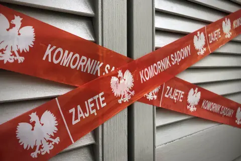Czerwona taśma z orłem i napisem "KOMORNIK SĄDOWY ZAJĘTE" krzyżuje się na tle szarej, żaluzjowej powierzchni. To znak, że nastąpiło zajęcie komornicze bez wezwania do zapłaty.