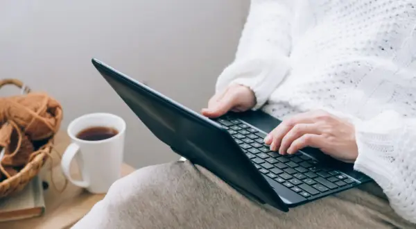 Osoba w białym swetrze uczy się, jak czytać raport BIK, pracując na laptopie przy kubku kawy i koszyku z włóczką.