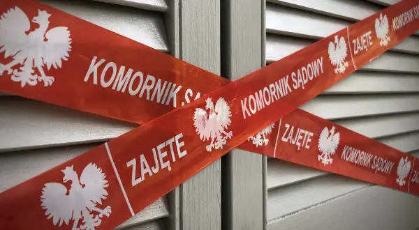 Czerwona taśma z orłem i napisem "KOMORNIK SĄDOWY ZAJĘTE" krzyżuje się na tle szarej, żaluzjowej powierzchni. To znak, że nastąpiło zajęcie komornicze bez wezwania do zapłaty.