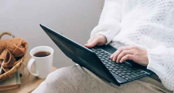 Osoba w białym swetrze uczy się, jak czytać raport BIK, pracując na laptopie przy kubku kawy i koszyku z włóczką.