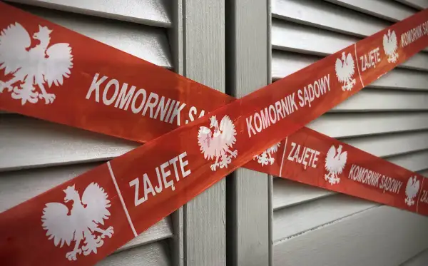 Czerwona taśma z orłem i napisem "KOMORNIK SĄDOWY ZAJĘTE" krzyżuje się na tle szarej, żaluzjowej powierzchni. To znak, że nastąpiło zajęcie komornicze bez wezwania do zapłaty.